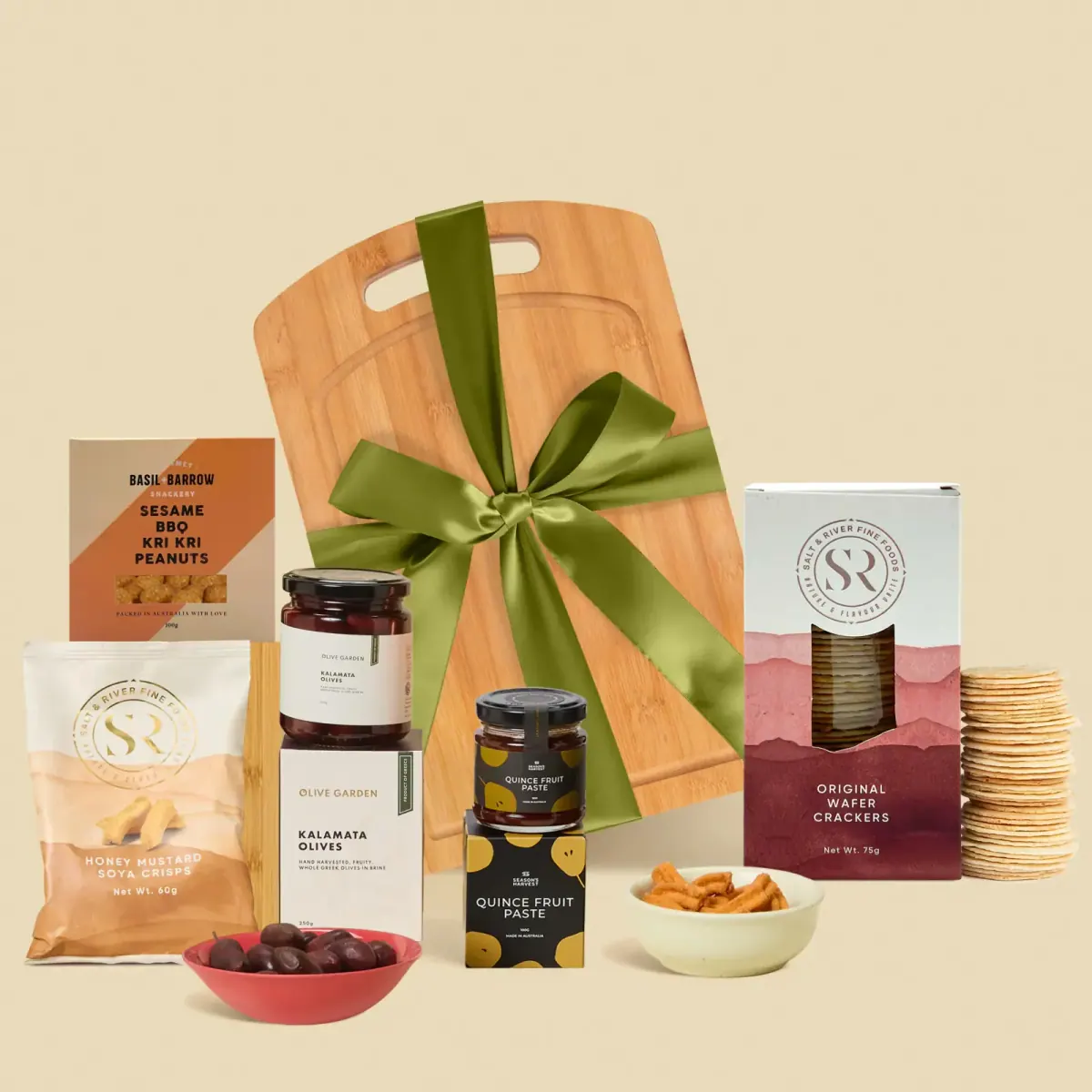A bamboo cheeseboard gift tied with a green ribbon, surrounded by kalamata olives, quince fruit paste, wafer crackers and soya crisps