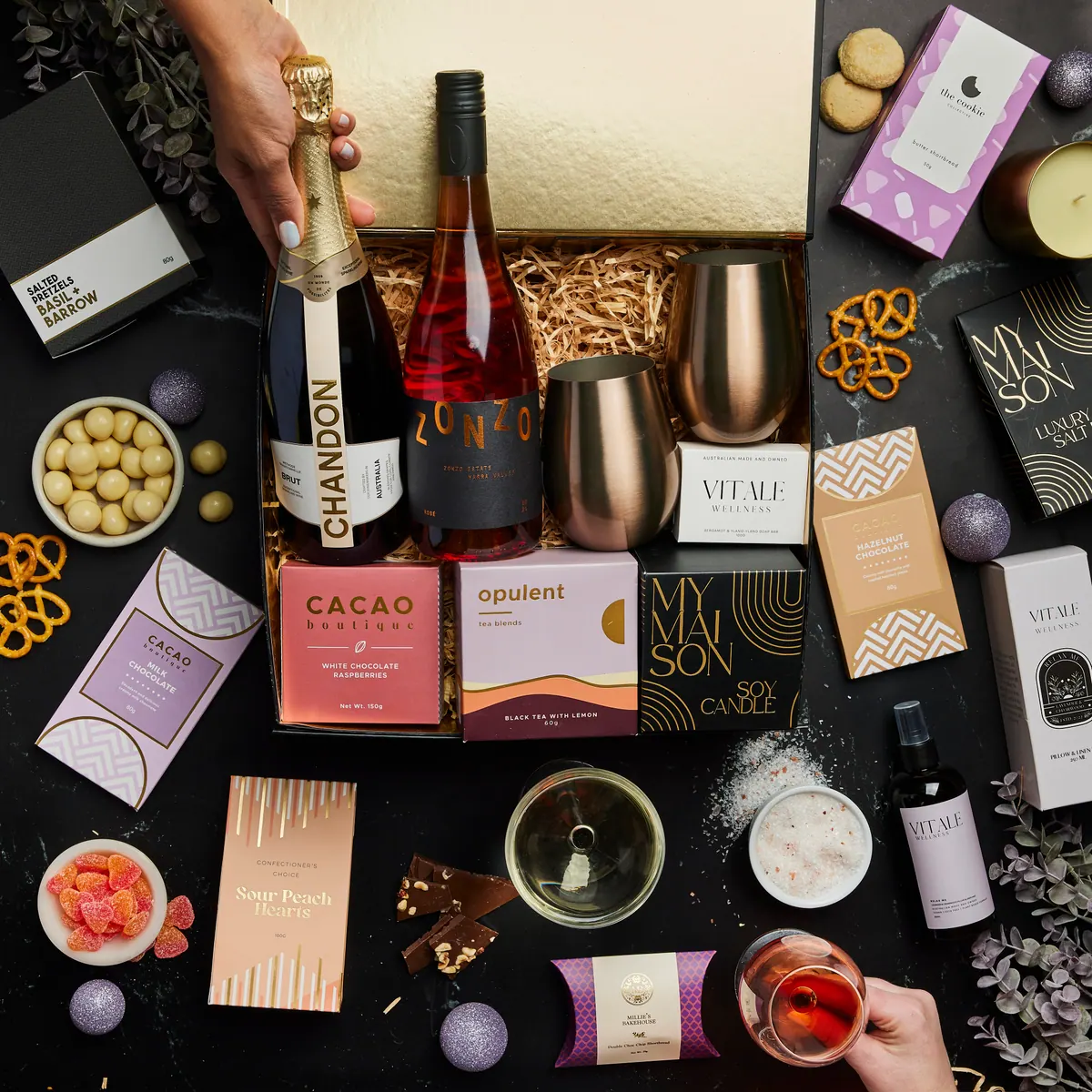 An open settlement gift hamper with Chandon sparkling wine, Zonzo rosé, two rose-gold tumblers, chocolate, tea, shortbread and a soy candle, laid out on a dark bench